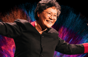 Maestra Mei-Ann Chen in a black suit against a bright pink and blue fireworks background.