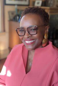 A headshot of Wendy Lewis wearing a pink blouse and chunky gold earrings.